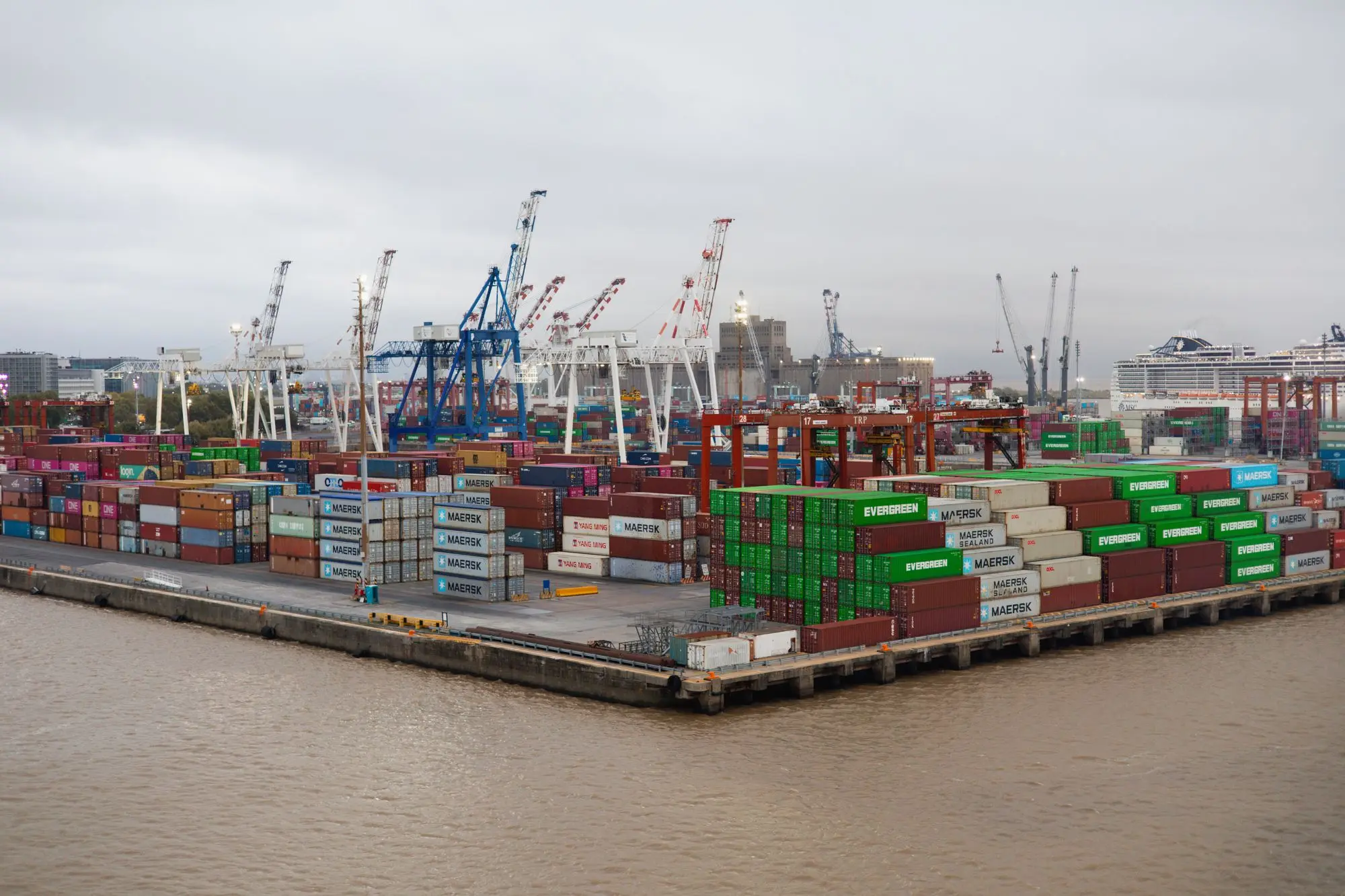 Stacked cargo containers and cranes at a working port under overcast sky.